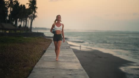 Attractive-blond-woman-holds-yoga-mat-walking-along-shore-in-Bali,-dusk