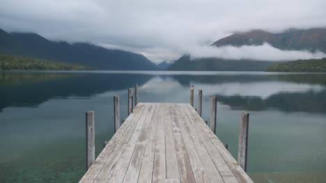 Static-shot-of-the-iconic-Rotoiti-lake-wooden-jetty-New-Zealand-on-a-cloudy-early-morning-in-4K-with-mountains-reflection-in-crystal-clear-water.