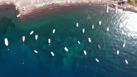 Aerial-top-down-view-descending-above-many-fishing-boats-in-the-Mediterranean-Sea-with-crytal-clear-blue-water-and-beach-in-Santorini,-Greece