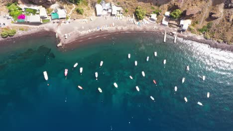 Aerial-top-down-view-descending-on-many-fishing-boats-in-the-Mediterranean-Sea-with-crytal-clear-blue-water-and-beach-in-Santorini,-Greece