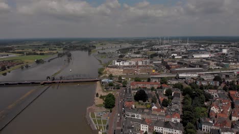 Aerial-backwards-movement-showing-the-countenance-cityscape-of-Zutphen,-The-Netherlands,-with-traffic-passing-by-on-the-boulevard-during-high-water-levels-of-the-river-IJssel-with-flooded-floodplains