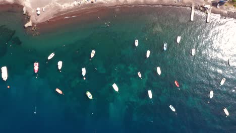 Aerial-top-down-view-flying-sideways-across-fishing-boats-in-the-Mediterranean-Sea-with-crytal-clear-blue-water-and-beach-in-Santorini,-Greece