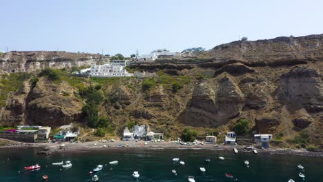 Aerial-static-view-of-Greek-Island,-rocky-cliff-with-villas-and-fishing-boats-in-the-Mediterranean-Sea-on-beach-in-Santorini,-Greece
