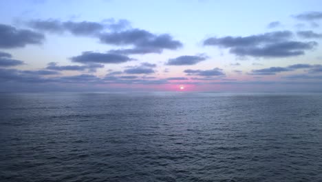 Low-light-scenic-sunset-above-vast-open-ocean-with-several-clouds-in-sky,-aerial