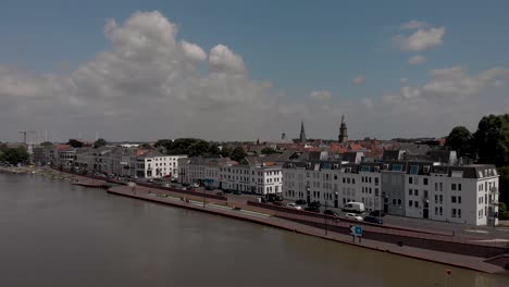 Aerial-approach-of-countenance-cityscape-of-Zutphen,-The-Netherlands,-with-traffic-passing-by-on-the-boulevard-with-white-facades-during-high-water-level-of-the-river-IJssel-passing-in-front