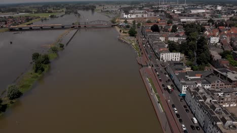 Aerial-descend-and-sideways-pan-showing-the-countenance-cityscape-of-Zutphen,-The-Netherlands,-with-traffic-passing-by-on-the-boulevard-during-high-water-levels-of-the-river-IJssel-filling-floodplains