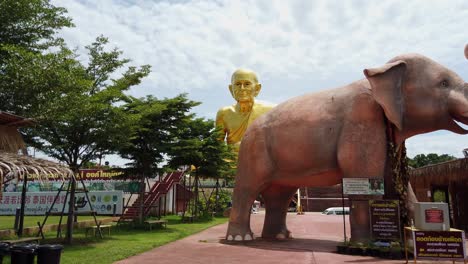 Schwenkaufnahme,-Die-Die-Leuchtend-Goldene-Mönchsstatue-Luang-Pu-Thuat-Im-Hintergrund-Und-Eine-Riesige-Elefantenskulptur-Im-Vordergrund-Im-Buddhistischen-Park-In-Phuttha-Utthayan-Maharat-Ayutthaya-Thailand-Zeigt