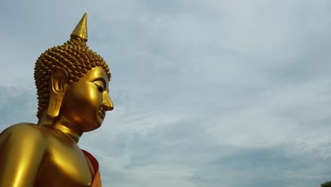 Close-up-dolly-out-profile-shot-of-a-majestic-divine-Buddhist-god,-the-lord-Gautama-Buddha-golden-statue-on-a-windy-day-with-overcast-grey-cloudy-sky-background-in-Thailand-Southeast-Asia