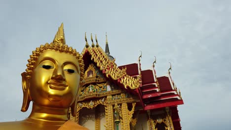 Dolly-out-shot-of-a-golden-Buddha-statue-and-Thai-Buddhist-Temple-with-ornate-style-of-high-gables,-stepped-out-roofs-and-elaborate-finials-in-an-overcast-weather-in-Thailand-Asia