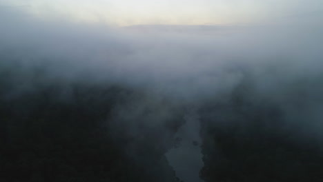 Aerial-drone-pushes-from-steep-river-valley-into-dense-fog-early-morning,-4K