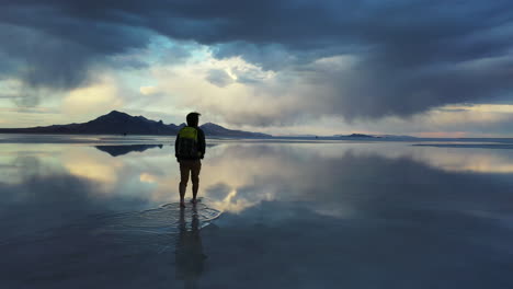 Silhouette-of-Man-With-Backpack-Walking-in-Surreal-Landscape-of-Salt-Flats-With-Stormy-Clouds-and-Sky-Mirror-Reflection-on-Water,-Cinematic-Approaching-View
