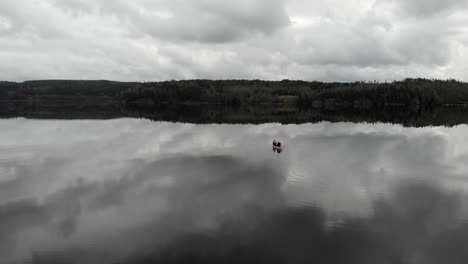 Dolly-in-descending-aerial-shot-of-two-people-paddling-gently-on-a-white-canoe-while-creating-small-ripples-all-over-a-calm-reflective-lake-towards-land-on-a-grey-cloudy-day