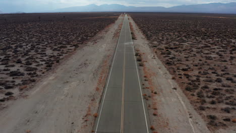 An-empty-and-lonely-road-through-the-heat-of-the-Mojave-Desert---tilt-up-aerial-view-with-mountains-beyond-the-flat-basin
