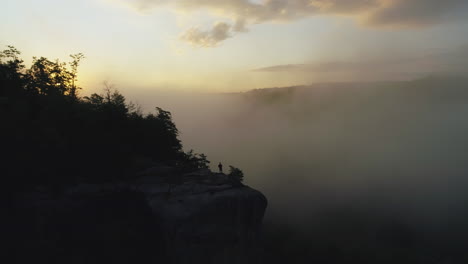 Aerial-passing-by-silhouette-on-foggy-cliff-overlook-during-sunrise,-4K