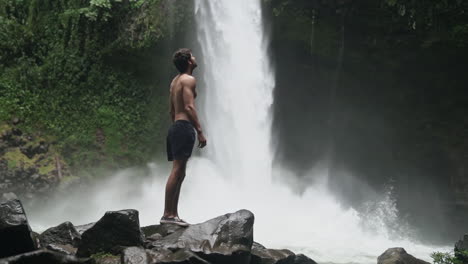 Shirtless-man-looks-up-to-top-of-La-Fortuna-waterfall-Costa-Rica,-slow-motion