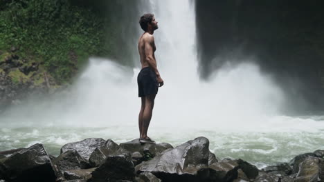 Man-looks-around-as-jungle-waterfall-crashes-down-behind-him,-slow-motion