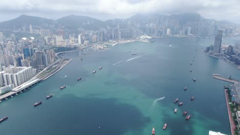 Convoy-of-local-Fishing-boats-causing-in-Hong-Kong-Victoria-bay,-with-city-skyline-in-the-horizon,-Aerial-view