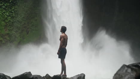 Man-in-front-of-roaring-waterfall-in-rain-forest-turns-to-look-up-at-cascade
