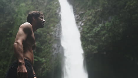 Low-angle-of-shirtless-man-looking-up-at-rushing-waterfall-in-green-rain-forest