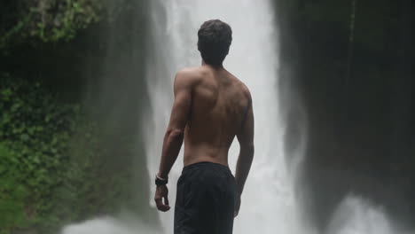 Man-turns-and-looks-at-rushing-La-Fortuna-waterfall-Costa-Rica,-slow-motion
