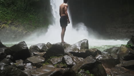 Man-stands-in-front-of-powerful-rain-forest-waterfall-with-mist-blowing-off