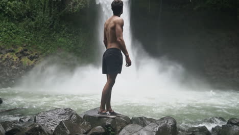 Pushing-in-as-man-turns-to-gaze-at-roaring-jungle-waterfall,-slow-motion