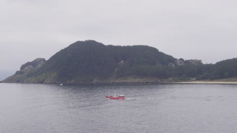 A-Red-Boat-Passing-by-A-Moody-and-Mysterious-Uninhabited-Island-in-CIes-Islands-Chain,-Spain