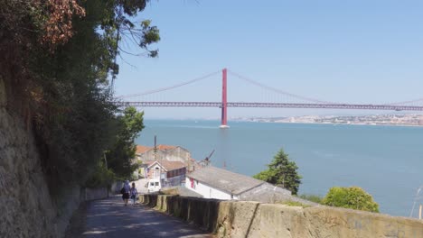 Ein-Paar,-Das-Die-Straße-Zu-Einem-Fischerdorf-Auf-Der-Anderen-Seite-Des-Flusses-In-Lissabon-Hinuntergeht,-Mit-Der-Brücke-25-De-Abril-Im-Hintergrund