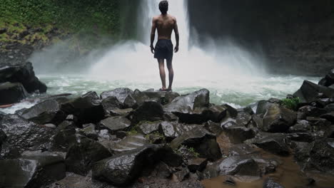 Panning-up-to-reveal-man-looking-up-at-thundering-jungle-waterfall,-slow-motion