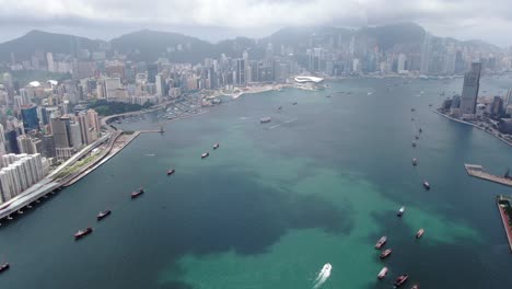 Convoy-of-local-Fishing-boats-causing-in-Hong-Kong-Victoria-bay,-with-city-skyline-in-the-horizon,-Aerial-view