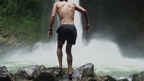 Man-walks-over-rocks-towards-roaring-La-Fortuna-waterfall-Costa-Rica,-slow-motion