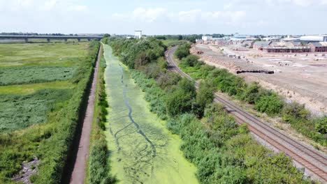Green-algae-covered-countryside-canal-alongside-new-housing-build-site-aerial-view-pan-right