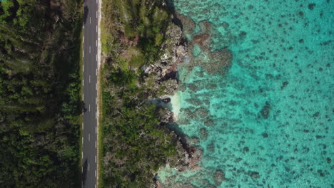 Bird's-eye-view-of-desert-road-along-rugged-shoreline,-crystal-water-in-Loyalty-islands,-New-Caledonia