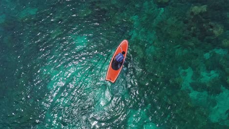 Top-View-Of-A-Man-Sitting-On-Paddle-Board-And-Paddling-Over-Clear-Blue-Lagoon-With-Beautiful-Coral-Reef-Underwater