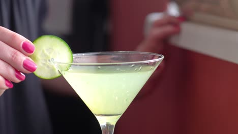 Cucumber-Wheel-Slice-Garnish-is-Placed-on-a-Martini-Glass-Filled-with-a-Green-Cocktail-or-Mocktail-Drink-Closeup,-Alcohol-Mixed-Drink-Made-by-Woman-Bartender-with-Pink-Nailpolish-Hands