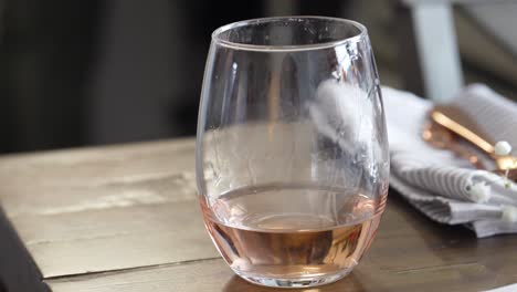 Woman-Picks-Up-Stemless-Wine-Glass-of-Rose-Half-Full-from-Wooden-Table,-Closeup-of-Hand-Wearing-Pink-Nail-Polish-and-Kitchen-Table-Napkin-and-Silverware-in-Background,-White-and-Golden-Red-Color