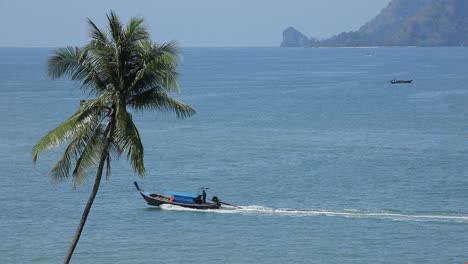 Longtail-boat-in-Krabi,-Thailand-passes-by-a-palm-tree-at-Ao-Nang-beach-with-island-sin-the-backround