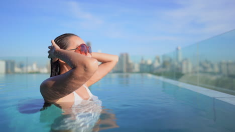 Young-asian-woman-standing-half-submerged-in-the-swimming-pool-on-a-rooftop,-rising-her-arms-and-touching-her-long-wet-hair-with-blurred-cityscape-in-the-backgroung