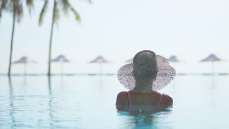 Eine-Frau-Mit-Dem-Rücken-Zur-Kamera-Im-Brusthohen-Poolwasser-Blickt-Auf-Die-Sonnenschirme-Und-Das-Meer