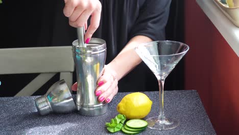 Bartender-Finishes-Muddling-Cocktail-Ingredients-in-a-Silver-Cobbler-Shaker-for-a-Mixed-Drink-in-a-Martini-Glass-with-Lemon,-mint-leaves-and-Cucumber-Slices,-Closeup-of-Woman’s-Hands-on-Granite-Table