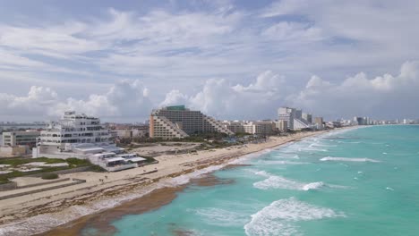 Turquoise-waves-breaking-on-sandy-shore-in-front-of-luxurious-beach-resorts,-Cancun,-Mexico