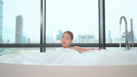 Beautiful-girl-relaxing-in-a-bathtube-with-white-foam-covering-her-body,-with-cityscape-seen-through-the-window
