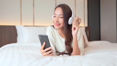 Schöne-Glücklich-Lächelnde-Thailändische-Frau,-Die-Mit-Kopfhörern-Auf-Dem-Bett-Liegt-Und-Musik-Hört,-Während-Sie-Das-Handy-Im-Hotelzimmer-Hält