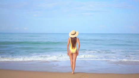 A-young-healthy-woman-in-a-yellow-bathing-suit-with-her-back-to-the-camera-walks-slowly-into-the-incoming-surf