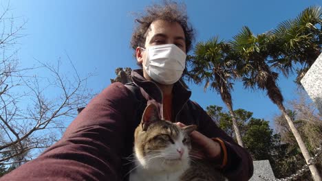 Male-vlogger-holding-selfie-camera-petting-cat-while-wearing-facemask