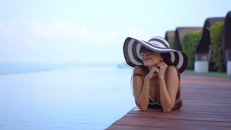 Asiatische-Lächelnde-Frau-Mit-Großem-Gestreiftem-Hut-Und-Vintage-Sonnenbrille-Beim-Sonnenbaden-Am-Pool