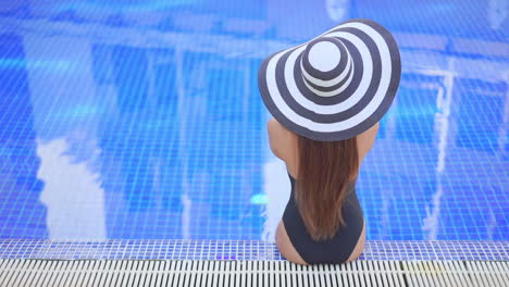 Back-view-of-a-female-tourist-sitting-by-the-swimming-pool-soaking-her-feet,-wearing-black-one-piece-swimwear-and-big-black-and-white-floppy-hat