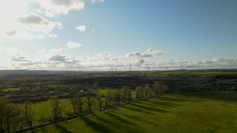 Aerial-drone-flies-over-green-fields-and-line-of-trees-in-spring,-Windmill-farm-factory-on-background-in-Puck,-Poland-on-sunny-and-cloudy-day