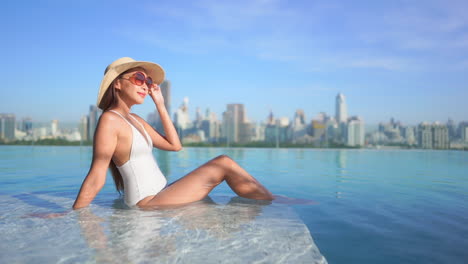 With-a-modern-skyline-in-the-background,-an-attractive-woman-in-a-bathing-suit-sits-in-the-shallow-end-of-an-infinity-edge-pool-enjoying-the-sun
