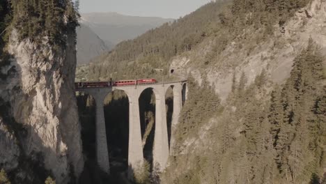 Train-cross-landwasser-viaduct-tunnels-between-mountains-covered-by-forests,-summer-season,-aerial-dolly-in